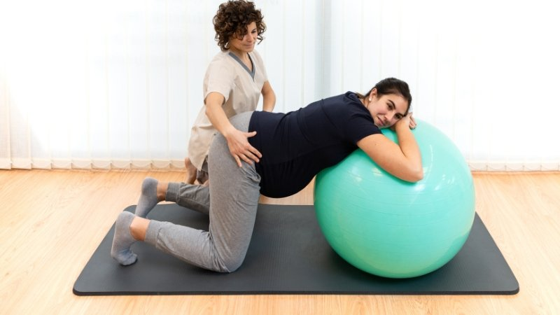 Pregnant woman exercising on medicine ball with the help of physiotherapist